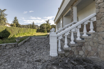 Stairs and exterior railings of a single-family home with white prefabricated balustrades on the façade with stones and floors of the same material and perimeter fence with hedges