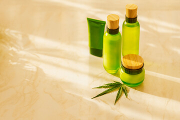 Green cosmetic bottles with wooden caps and hemp leaf on marble surface.