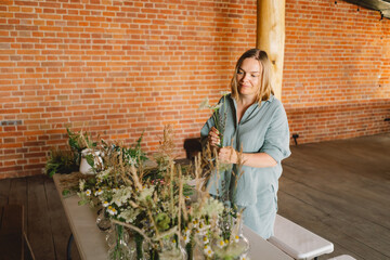 A woman stands at a wooden table, artfully arranging assorted green plants and branches in a...