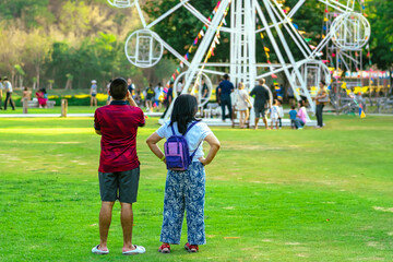 Back view of Asian tourists enjoy outdoor theme park together with beautiful white ferris wheel...