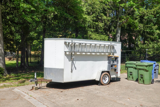 White contractor's supply trailer with attached ladder in a driveway near trash cans