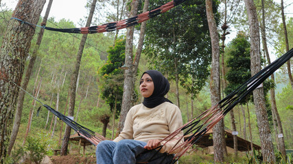 Pensive smiling young woman with headscarf resting in comfortable hammock at green garden.