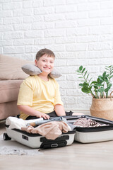 Happy smiling boy with yellow shirt ready for vacation. Happy child packs clothes into a suitcase for travel. Tourist, joy of holiday. Kid at home, preparing for flying. Modern and cozy interior.
