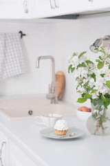 Spring morning in light kitchen: bouquet of white flowers, cake, glass of green tea, apples in white bowl.