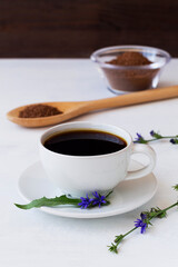close-up of hot chicory drink in white cup decorated blue chicory flowers with Ground chicory root on a wooden spoon on white background. healthy. coffee substitute. copy space. vertical