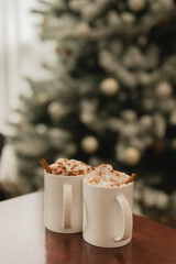Two mugs with hot chocolate with whipped cream and cinnamon powder & cinnamon sticks against a Christmas tree background
