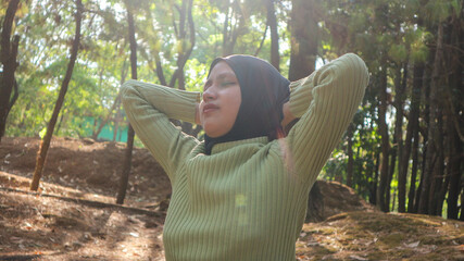 muslim woman with hands behind head and eyes closed breathing fresh air in middle of pinewood forest