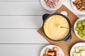 Fondue pot with tasty melted cheese, fork and different snacks on white wooden table, flat lay....