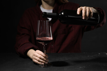 Alcohol addiction. Woman pouring red wine from bottle into glass at dark textured table, closeup