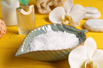 Natural sea salt in bowl and other spa products on yellow wooden table, closeup