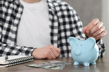 Financial savings. Man putting coin into piggy bank at wooden table, closeup