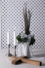 Burning candles, bouquet with willow branches and cross on white wooden table