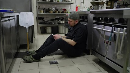 A tired male chef in a black uniform sits on the floor and checks social networks using his phone during a short break in the middle of a busy working day in the kitchen of a restaurant - Powered by Adobe