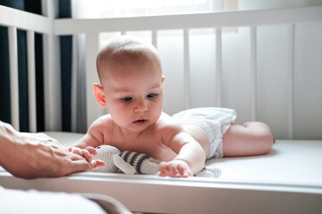 A charming and cute infant baby lies in a crib on white linen in a diaper and looks around while...