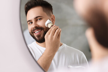 Handsome young man shaving beard near mirror in bathroom
