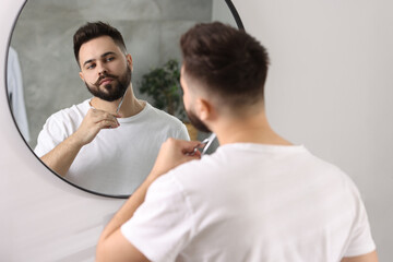 Handsome young man trimming beard with scissors near mirror in bathroom