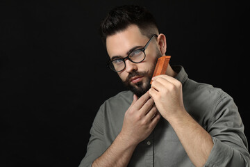 Handsome young man combing beard on black background. Space for text