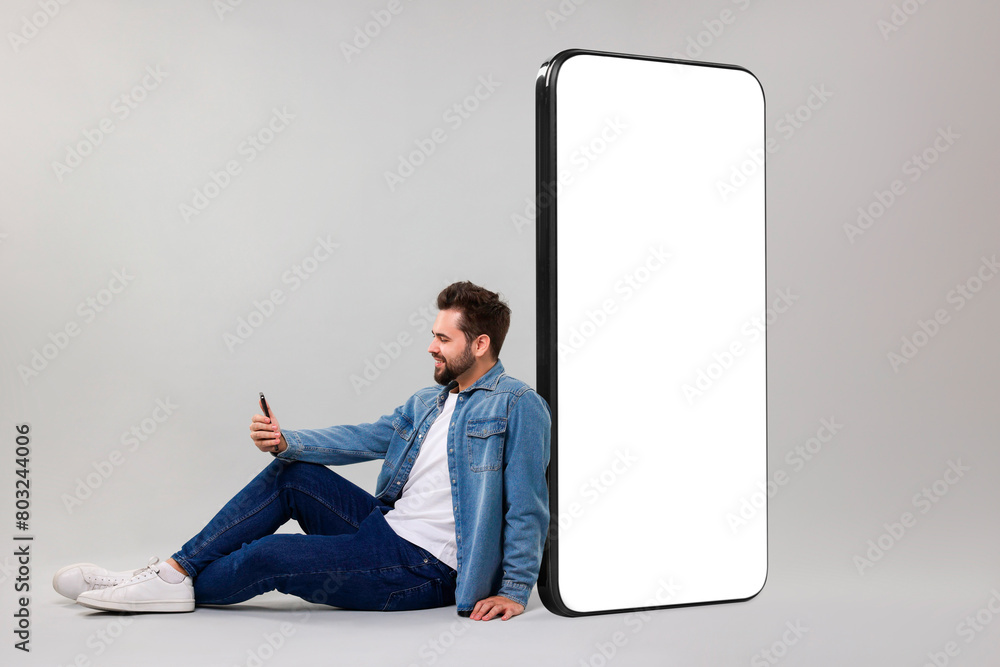 Aufkleber man with mobile phone sitting near huge device with empty screen on grey background. mockup for desi