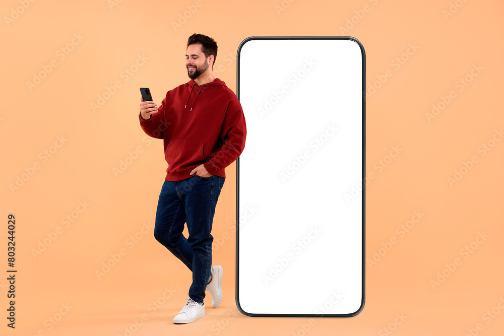 Aufkleber man with mobile phone standing near huge device with empty screen on dark beige background. mockup f