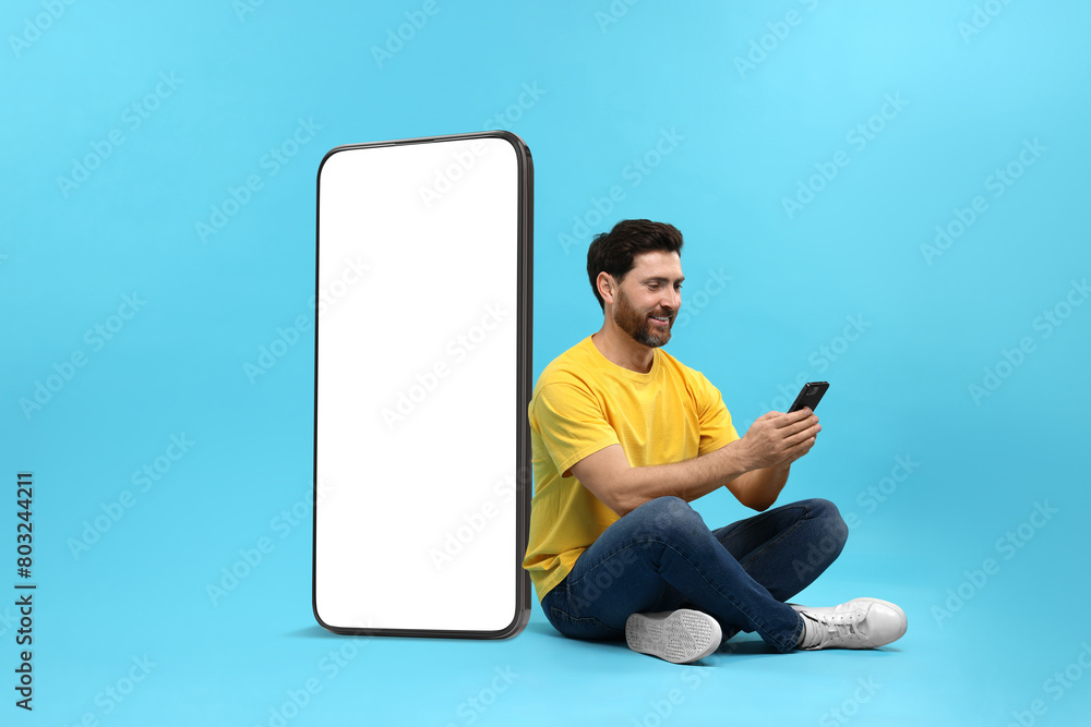 Poster man with mobile phone sitting near huge device with empty screen on light blue background. mockup fo