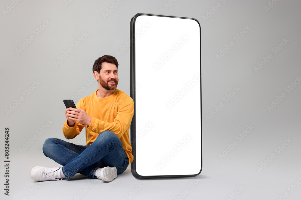 Poster man with mobile phone sitting near huge device with empty screen on grey background. mockup for desi