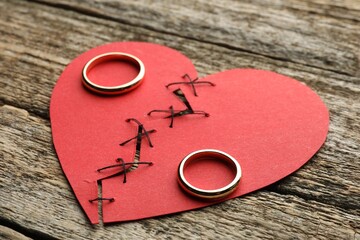 Broken heart. Torn red paper heart sewed with thread and wedding rings on wooden table, closeup