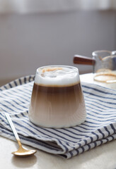 A clear glass of iced latte coffee layer with mousse, spoon and pot in background. It's a beverage...