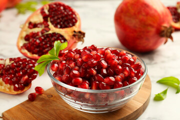 Ripe juicy pomegranate grains in bowl and green leaves on white table