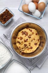 Dough with raisins in bowl and ingredients on white wooden table, flat lay