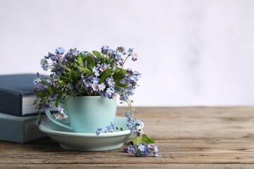 Beautiful forget-me-not flowers in cup, saucer and books on wooden table against light background, closeup. Space for text