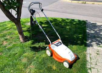 White-orange manual electric lawn mower on a lush green lawn near a tree in the city on a sunny spring day