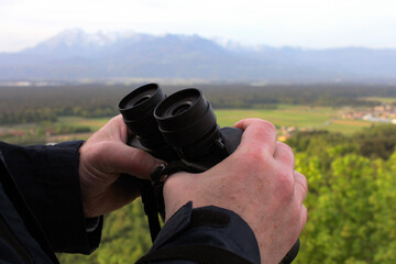 A man holds binoculars with both hands against the backdrop of the Alps and forest
