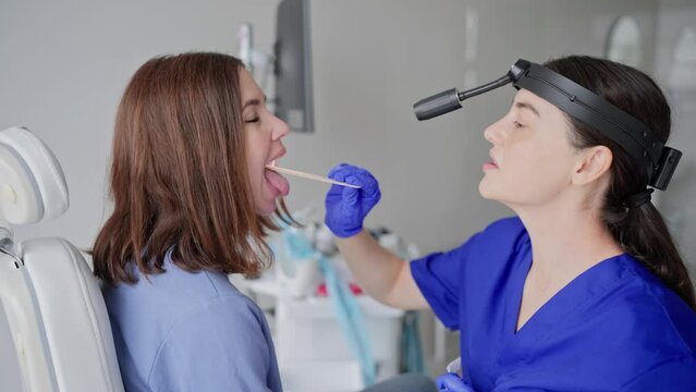 Close-up of a confident young brunette girl at an appointment with an ENT doctor in a modern clinic. A brunette doctor girl in a blue uniform puts a special stick in the patients mouth to check the