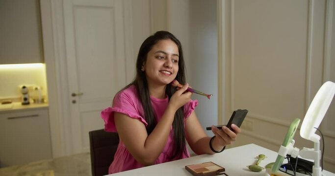 A happy Indian brunette girl in a pink dress sits in front of the lighting and her green smartphone while blogging online about how to properly apply cosmetics and do Makeup in a modern apartment