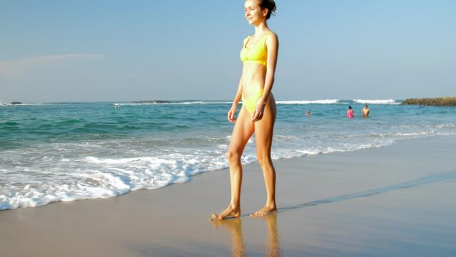 Slender model wearing bikini strolls along damp coastline of ocean slow motion. Woman smiles as enjoys time on peaceful beach far from urban clamor