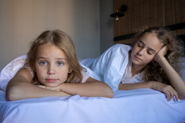Portrait of two blonde little girls sisters on a large and cozy white bed with a dissatisfied and...
