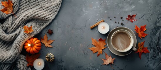 Autumn comfort at home: An aerial image featuring a cup of steaming coffee, a cozy blanket, pumpkin candles, maple leaves, and fragrant spices on a grey backdrop with space for text.