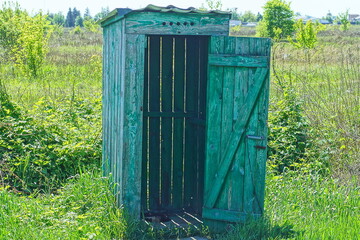 wooden with green boards, public, homemade, primitive, with a roof and an open door, the toilet stands on the street on green grass during the day