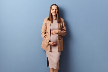 Brown haired pretty elegant pregnant woman in beautiful dress and jacket posing isolated over blue...