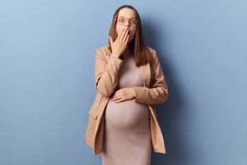 Shocked scared pregnant woman in beautiful dress and jacket posing isolated over blue background...