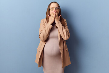 Astonished surprised pregnant woman in beautiful dress and jacket covering mouth with both hands...