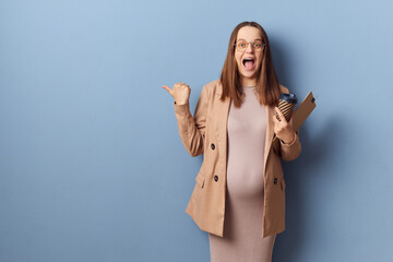 Excited pregnant woman in beautiful dress and jacket posing isolated over blue background showing...