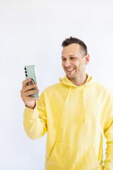 young smiling happy fun man 20s wear yellow hoodie hold in hand use mobile cell phone isolated on white background studio portrait. People lifestyle concept. 