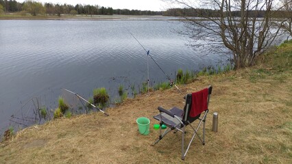 A folding fishing chair, landing net, a towel, bait boxes, a bucket with lure, a feeder rod for bream fishing mounted on stands next to bushes on the river bank. Flood. On the far bank is a forest