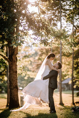 Valmiera, Latvia - August 19, 2023 -  a groom lifts his bride in a sunlit park, both smiling, with...