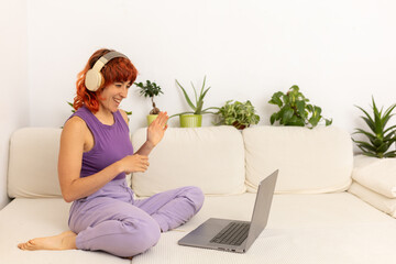 A woman is sitting on a couch with a laptop in front of her. She is wearing headphones and smiling....
