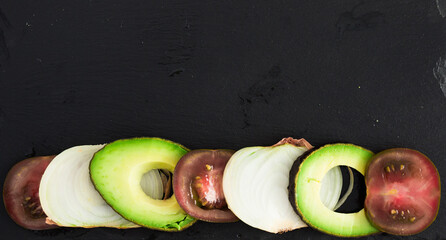 Chopped vegetables black tomatoes, avocado and onions against black stone board