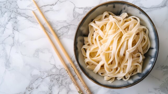 Dried udon nudles in a bowl closeup. Bunch of raw japanese vegan pasta from wheat flour and bamboo chopsticks over bowl against marble countertop.