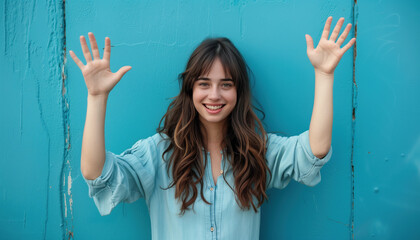 Happy young woman with brown hair in blue against a blue wall holding her hands up
