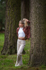 View of a young woman in a burgundy shirt, short white top, light jeans with blond hair. A girl stands among tall trees in a city park. Urban lifestyle concept, relaxing in nature